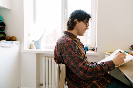 Téléchargez les photos : Homme blanc riant et écrivant des notes tout en étant assis dans la cuisine à la maison - en image libre de droit