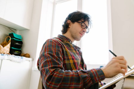 Téléchargez les photos : Homme blanc portant des lunettes écrivant des notes tout en étant assis dans la cuisine à la maison - en image libre de droit