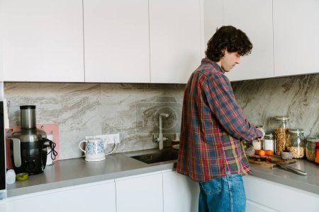 Téléchargez les photos : Homme blanc portant une chemise coupant des légumes pendant la cuisson du déjeuner dans la cuisine à la maison - en image libre de droit