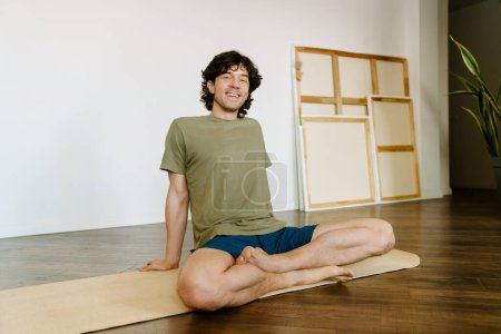 Téléchargez les photos : Homme blanc riant et assis sur le tapis pendant la pratique du yoga à la maison - en image libre de droit