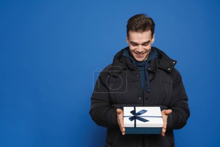 Foto de Joven hombre blanco sonriendo mientras posando con caja de regalo aislado sobre fondo azul - Imagen libre de derechos