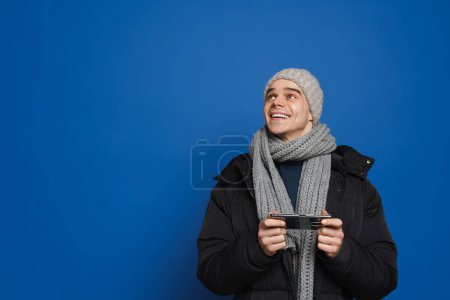 Téléchargez les photos : Jeune homme blanc portant des vêtements d'hiver à l'aide d'un téléphone cellulaire isolé sur fond bleu - en image libre de droit