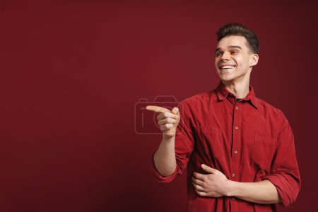 Téléchargez les photos : Jeune homme blanc portant chemise pointant doigt de côté et riant isolé sur fond rouge - en image libre de droit