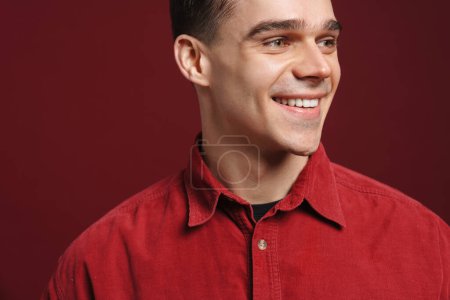 Foto de Joven hombre blanco vistiendo camisa sonriendo y mirando a un lado aislado sobre fondo rojo - Imagen libre de derechos