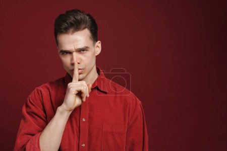 Téléchargez les photos : Jeune homme blanc fronçant les sourcils et montrant un geste de silence à la caméra isolé sur fond rouge - en image libre de droit