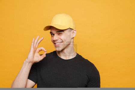 Foto de Joven hombre blanco con gorra guiñando el ojo y mostrando ok signo aislado sobre fondo amarillo - Imagen libre de derechos