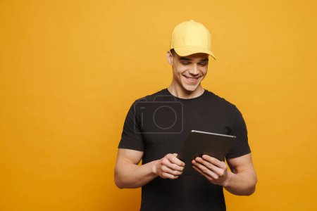 Foto de Joven hombre blanco con gorra sonriendo mientras usa tableta aislada sobre fondo amarillo - Imagen libre de derechos