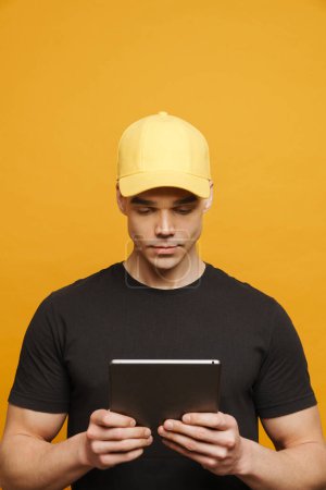 Foto de Hombre blanco joven usando la escritura de la tapa mientras que usa la computadora de la tableta aislada sobre fondo amarillo - Imagen libre de derechos