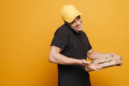 Téléchargez les photos : Jeune livreur parlant sur son téléphone portable tout en tenant des boîtes à pizza isolées sur fond jaune - en image libre de droit