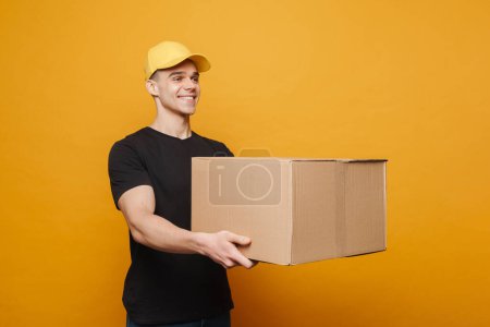 Téléchargez les photos : Jeune livreur souriant tout en posant avec boîte en carton isolé sur fond jaune - en image libre de droit