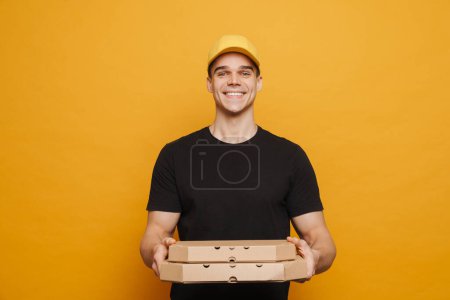 Téléchargez les photos : Jeune livreur souriant tout en posant avec des boîtes à pizza isolées sur fond jaune - en image libre de droit