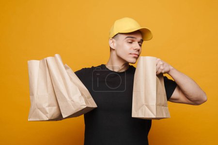 Téléchargez les photos : Jeune livreur portant une casquette posant avec des sacs en papier isolés sur fond jaune - en image libre de droit