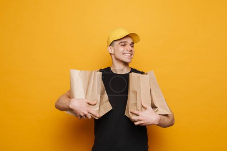 Foto de Joven repartidor sonriendo posando con bolsas de papel aisladas sobre fondo amarillo - Imagen libre de derechos