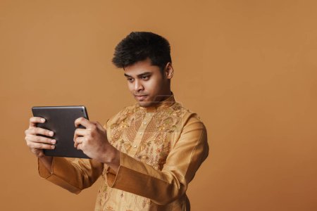 Téléchargez les photos : Jeune homme indien brune tenant et regardant tablette isolée sur fond jaune - en image libre de droit