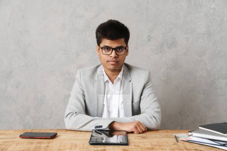 Foto de Joven morena empresario indio utilizando tableta y sentado en la mesa en la oficina sobre la pared gris - Imagen libre de derechos