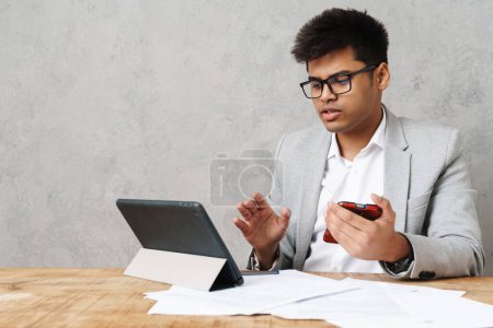 Foto de Joven empresario indio serio usando tableta y teléfono celular mientras trabaja en la oficina - Imagen libre de derechos
