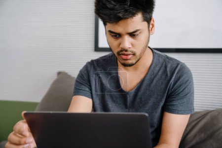 Téléchargez les photos : Jeune homme indien concentré regardant l'écran d'ordinateur portable assis sur le lit dans une chambre confortable à la maison - en image libre de droit