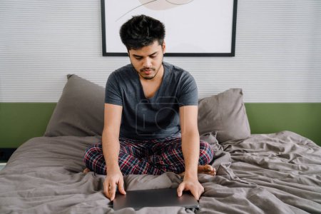 Téléchargez les photos : Jeune homme calme indien assis sur le lit avec ordinateur portable fermé à la maison - en image libre de droit
