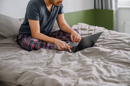 Téléchargez les photos : Jeune homme assis sur le lit et travaillant sur ordinateur portable dans une chambre confortable à la maison recadrée - en image libre de droit