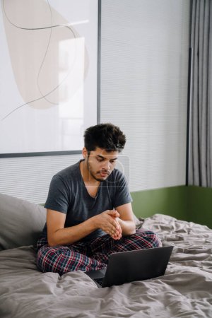 Téléchargez les photos : Jeune bel homme indien assis sur le lit avec les mains pliées et regardant ordinateur portable dans une chambre confortable à la maison - en image libre de droit