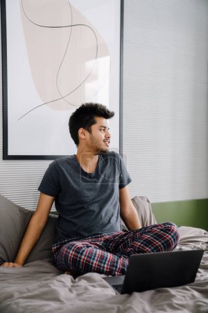 Téléchargez les photos : Jeune bel homme indien assis sur le lit en pose de lotus avec ordinateur portable et regardant de côté dans une chambre confortable à la maison - en image libre de droit