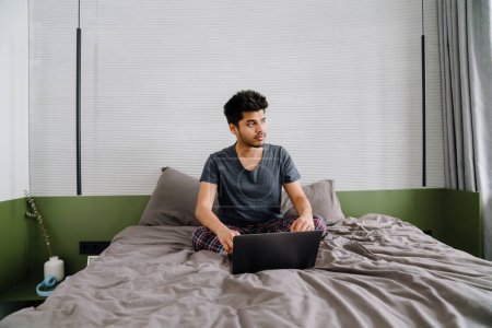 Photo for Young handsome indian man sitting on bed in lotus pose with laptop and looking aside in cozy bedroom at home - Royalty Free Image
