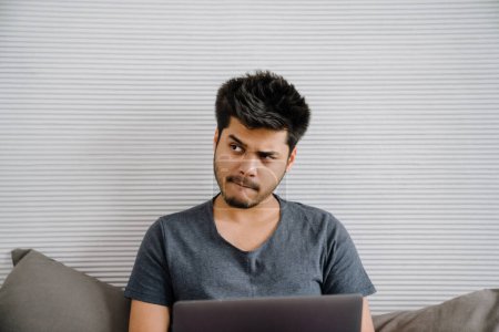 Téléchargez les photos : Jeune bel homme indien réfléchi avec ordinateur portable regardant de côté assis sur fond blanc rayé à la maison - en image libre de droit