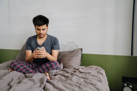 Foto de Joven indio con teléfono sentado en pose de loto en la cama en casa - Imagen libre de derechos