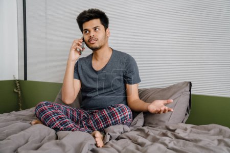 Téléchargez les photos : Jeune homme souriant indien parlant sur son téléphone assis dans la pose de lotus sur son lit - en image libre de droit
