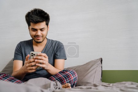 Photo for Young indian smiling man scrolling his phone sitting in lotus pose on his bed at home - Royalty Free Image