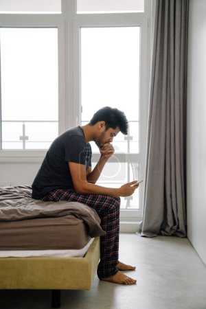 Foto de Joven hombre indio guapo sentado en la cama con teléfono y mirando en él en el acogedor dormitorio en casa - Imagen libre de derechos