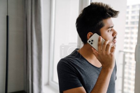 Téléchargez les photos : Jeune bel homme indien parlant sur son téléphone et regardant par la fenêtre à la maison - en image libre de droit
