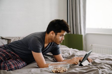 Photo for Young handsome indian man liying on bed working with tablet with bowl of snack in cozy bedroom athome - Royalty Free Image