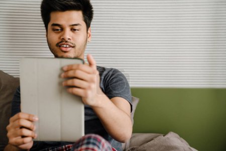 Téléchargez les photos : Jeune bel homme indien souriant tenant tablette regardant tout en étant assis à la maison - en image libre de droit