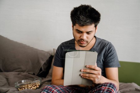 Téléchargez les photos : Jeune homme indien faisant défiler la tablette assis sur le lit avec bol de noix à la maison - en image libre de droit