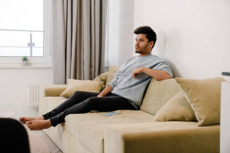 Téléchargez les photos : Jeune homme indien relaxant et assis sur le canapé dans une chambre confortable à la maison - en image libre de droit