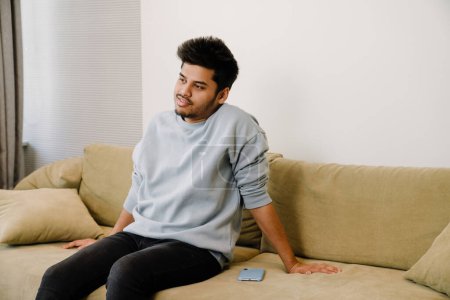 Téléchargez les photos : Jeune homme indien relaxant et assis sur le canapé à côté de son téléphone dans une chambre confortable à la maison - en image libre de droit