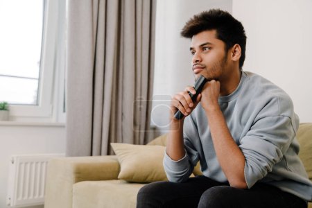 Photo for Young indian man sitting on sofa and watching tv at home - Royalty Free Image