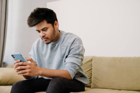 Photo for Young indian man sitting on sofa and scrolling news on his phone at home - Royalty Free Image