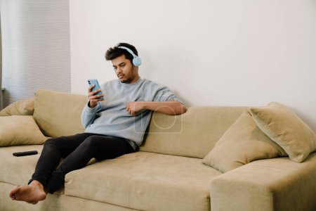 Téléchargez les photos : Jeune homme indien dans les écouteurs avec téléphone assis sur le canapé à la maison - en image libre de droit