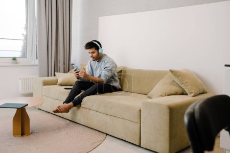 Téléchargez les photos : Jeune Indien en casque d'écoute tenant une vidéoconférence assis sur le canapé à la maison - en image libre de droit