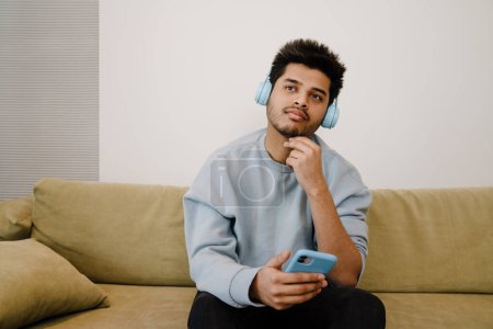 Téléchargez les photos : Jeune homme réfléchi indien dans les écouteurs écoute conférence assis sur le canapé à la maison - en image libre de droit