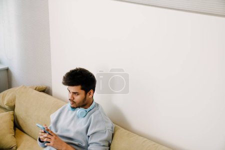 Téléchargez les photos : Jeune homme indien ennuyeux avec des écouteurs sur son cou tenant le téléphone et assis sur le canapé à la maison - en image libre de droit