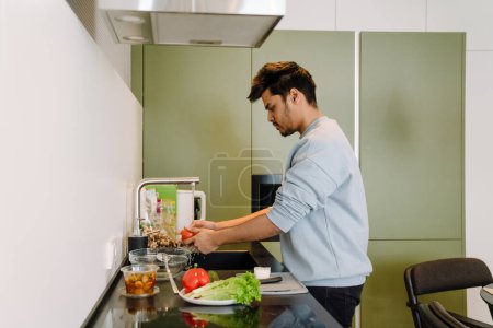 Téléchargez les photos : Jeune homme indien lavant des légumes pour cuisiner une salade sur une cuisine confortable à la maison - en image libre de droit