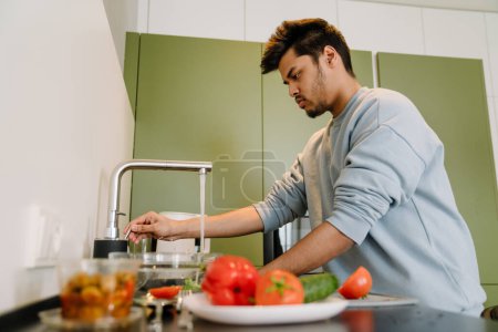 Téléchargez les photos : Homme indien adulte lavant des légumes pour cuisiner une salade dans une cuisine confortable à la maison - en image libre de droit