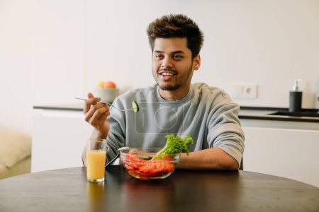 Téléchargez les photos : Jeune homme souriant indien mangeant une salade fraîche avec un verre de jus d'orange à la maison - en image libre de droit