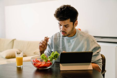 Téléchargez les photos : Jeune homme indien avec tablette mangeant une salade fraîche avec un verre de jus d'orange à la maison - en image libre de droit