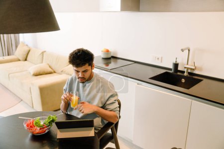 Photo for Young indian man looking on tablet while having lunch with bowl of fresh salad and glass of orange juice - Royalty Free Image