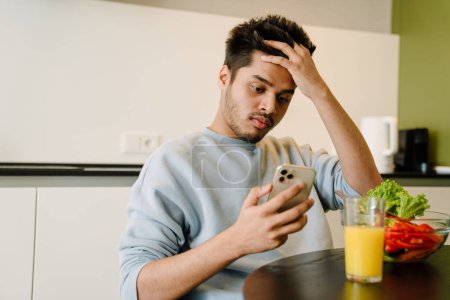 Téléchargez les photos : Jeune homme indien regardant au téléphone tout en déjeunant avec un bol de salade fraîche et un verre de jus d'orange - en image libre de droit