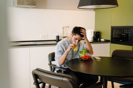 Photo for Young indian man propping his head by his hand sitting on kitchen with bowl of fresh salad and glass of orange juice - Royalty Free Image